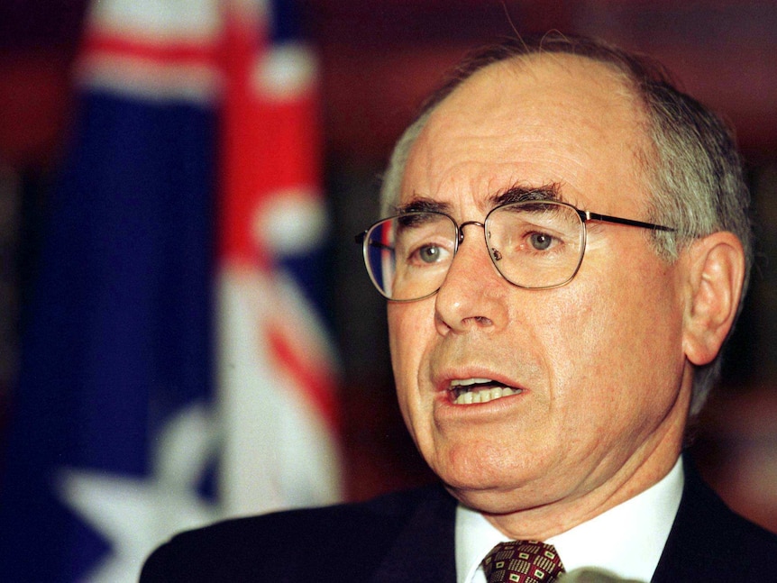 John Howard, with bushy dark eyebrows and his signature glasses, stands in front of an Australian flag.