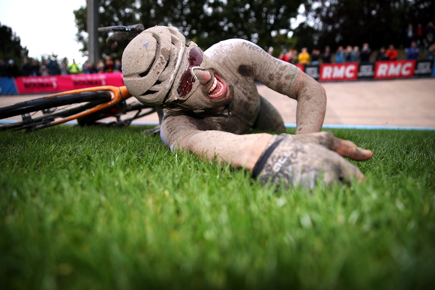 Sonny Colbrelli, completely covered in mud and still wearing his helmet, lays on grass and screams