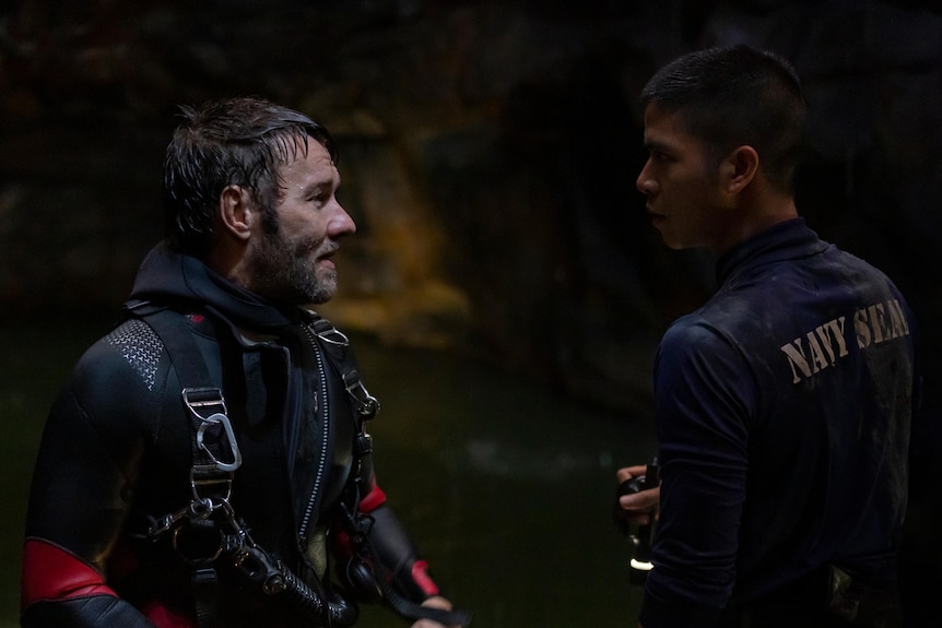 White man in wetsuit and with wet hair standing looking hopeless in front of Thai man in Navy Seal shirt and torch