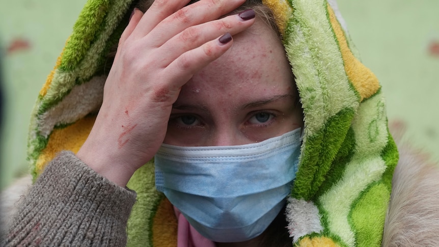 A woman wearing a face mask and hood raises a hand to her forehead.