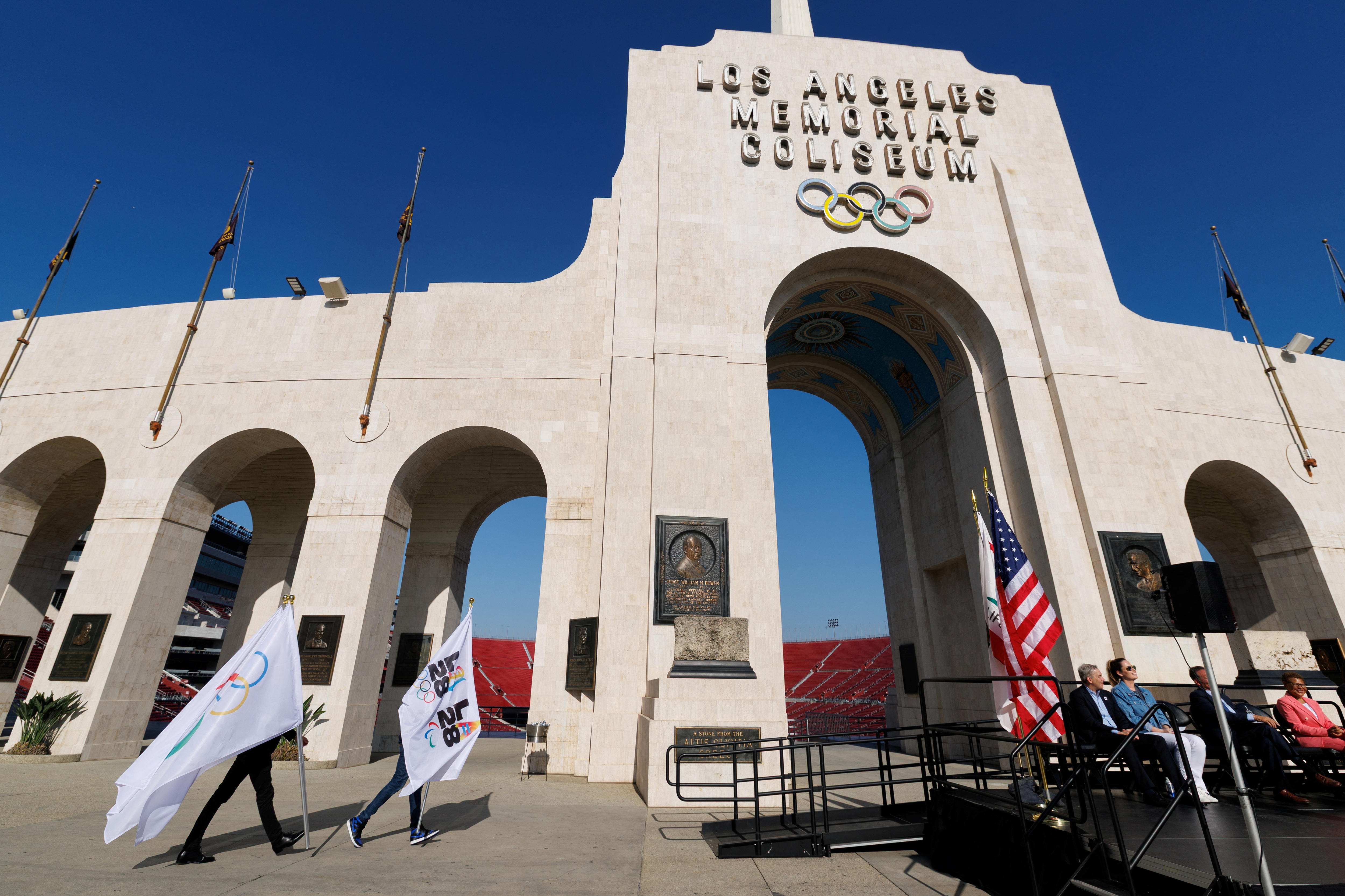 People carrying white LA28 Olympic flags alongside the sandstone Los Angeles Memorial Coliseum structure