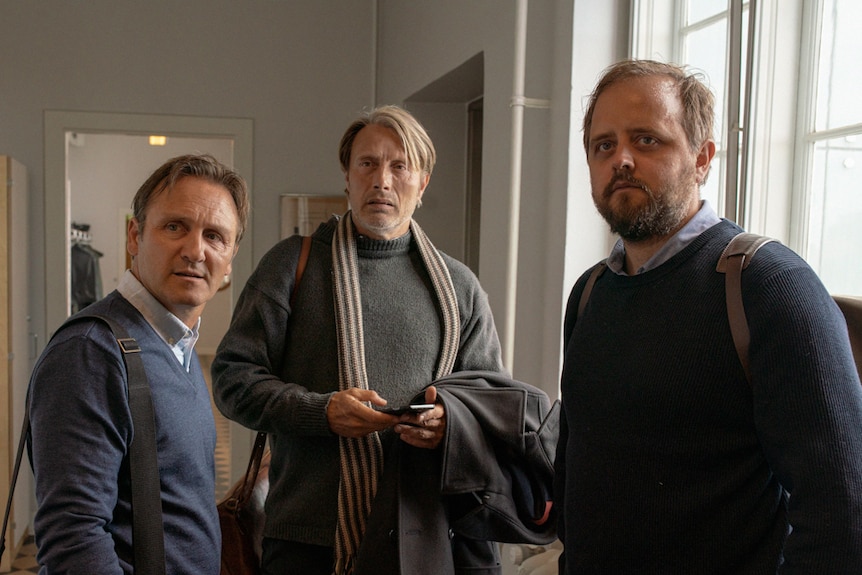 Interior shot of the three men standing together and looking in one direction as if concerned.