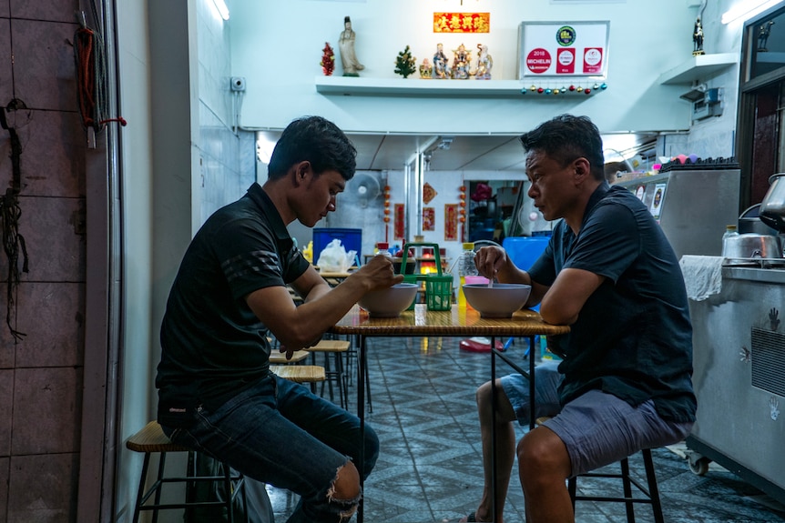 A young Laotian man and a 51-year-old Taiwanese man sit opposite each other eating, at a roadside shop.