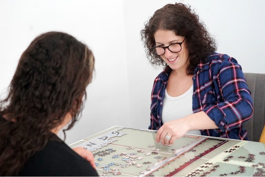 A woman sitting at a table, doing a puzzle, with another women who is seen from behind.