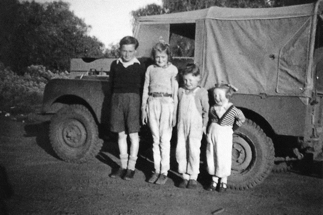 Tim Fischer, as a young boy wearing overalls, poses for a photo with his three young siblings in front of a vehicle.