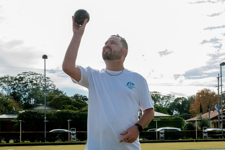 A man in his early thirties stands on a lawn bowls green. he is wearing a white shirt and holding a bowl like a tribute to god