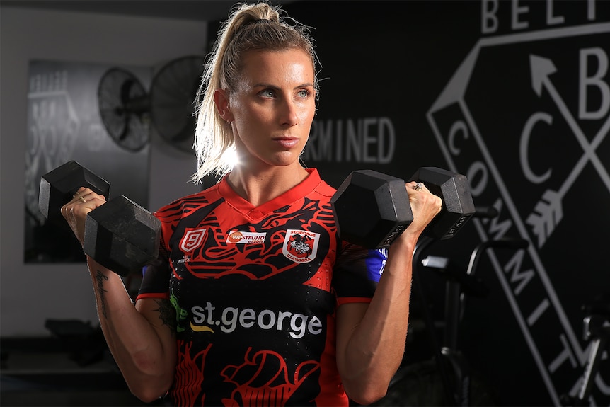 Sam Bremner holds up two dumbbells in a gym, wearing his St George Illawarra Dragons uniform.