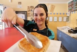 A girl smiling working at a bakery holding a pie to the camera with tongs.