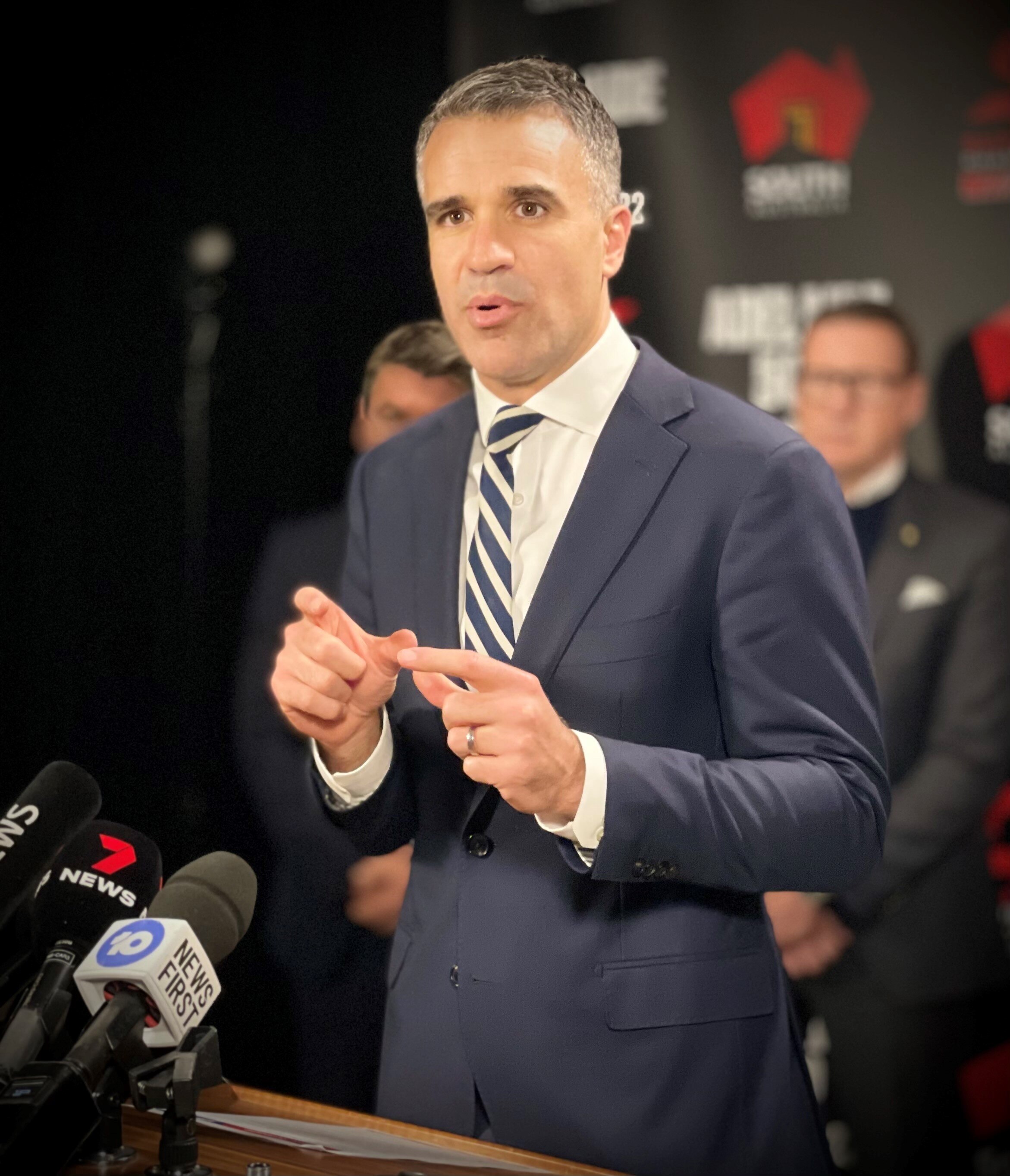 A man in a blue suit speaks into media microphones at a podium with a banner behind him with the SA logo