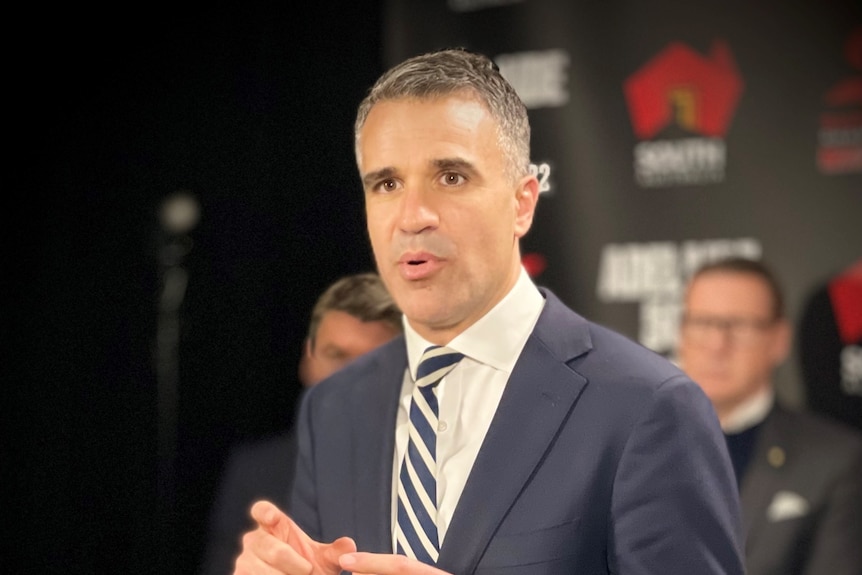 A man in a blue suit speaks into media microphones at a podium with a banner behind him with the SA logo