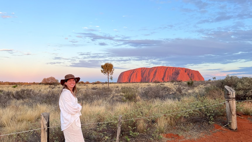 Woman wearing white standing on red dirt with brown vegetation with bright blue sky with bright red Uluru in background