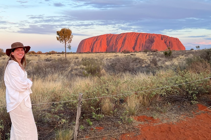 Woman wearing white standing on red dirt with brown vegetation with bright blue sky with bright red Uluru in background