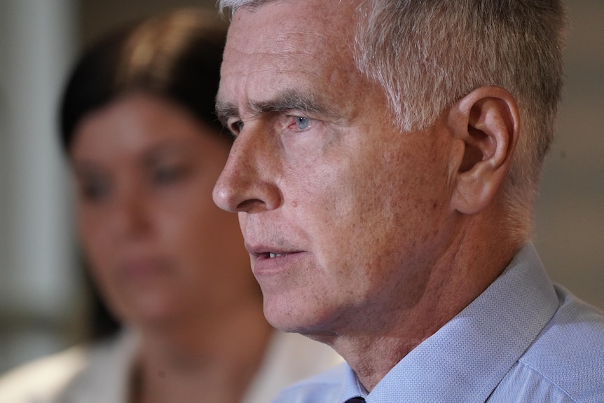 A man with a serious expression wearing a light blue shirt looks off camera.