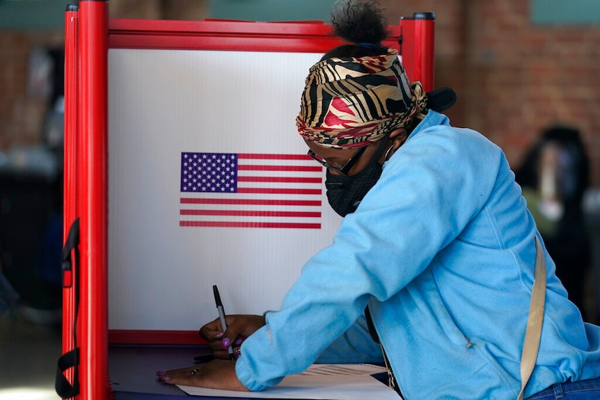 You view a woman of African American descent marking a ballot in a voting booth with a large American flag.