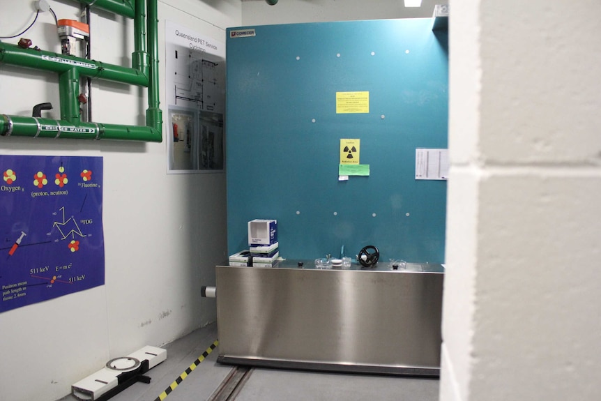 A white walled room with posters and green pipes on the walls and a teal coloured concrete door labelled with caution signs.