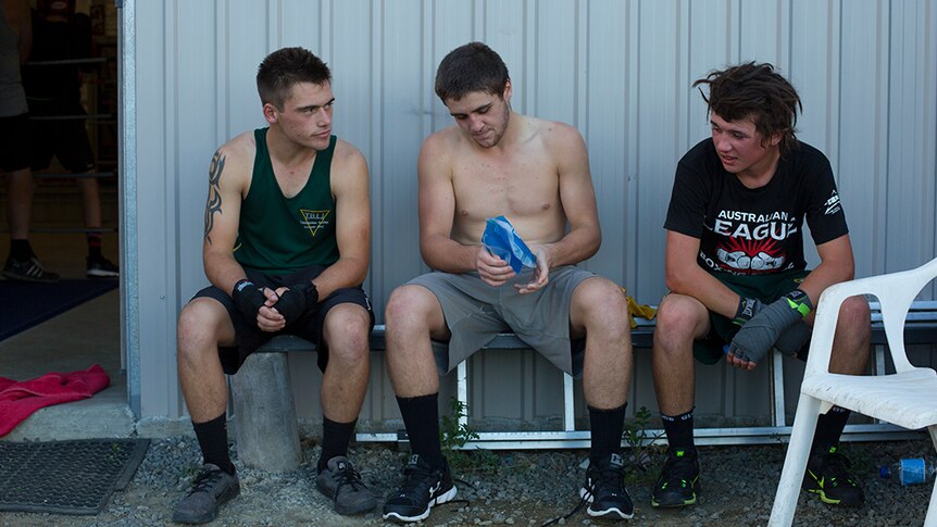 Three male boxers sit down to take a break outside the gym as the centred man applies an ice pack to his hand.