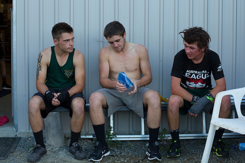 Three male boxers sit down to take a break outside the gym as the centred man applies an ice pack to his hand.