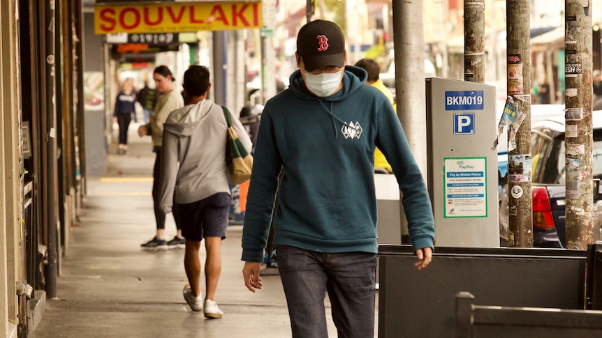 A man wearing jeans, a hoodie, baseball cap and white mask walks down a street.