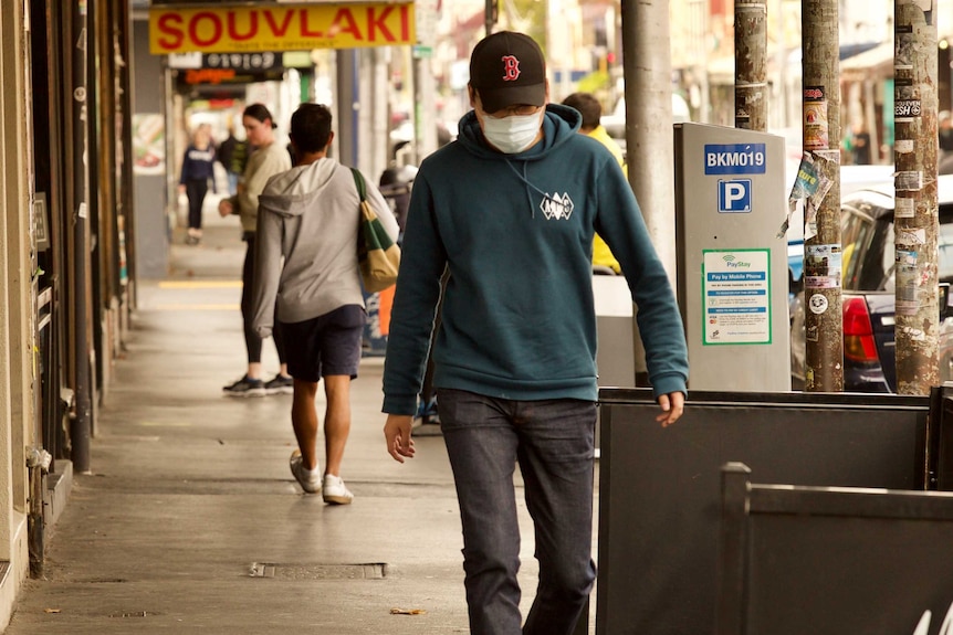 A man wearing jeans, a hoodie, baseball cap and white mask walks down a street.