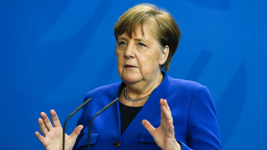 German Chancellor Angela Merkel gestures with both hands while wearing a blue blazer against a blue background.