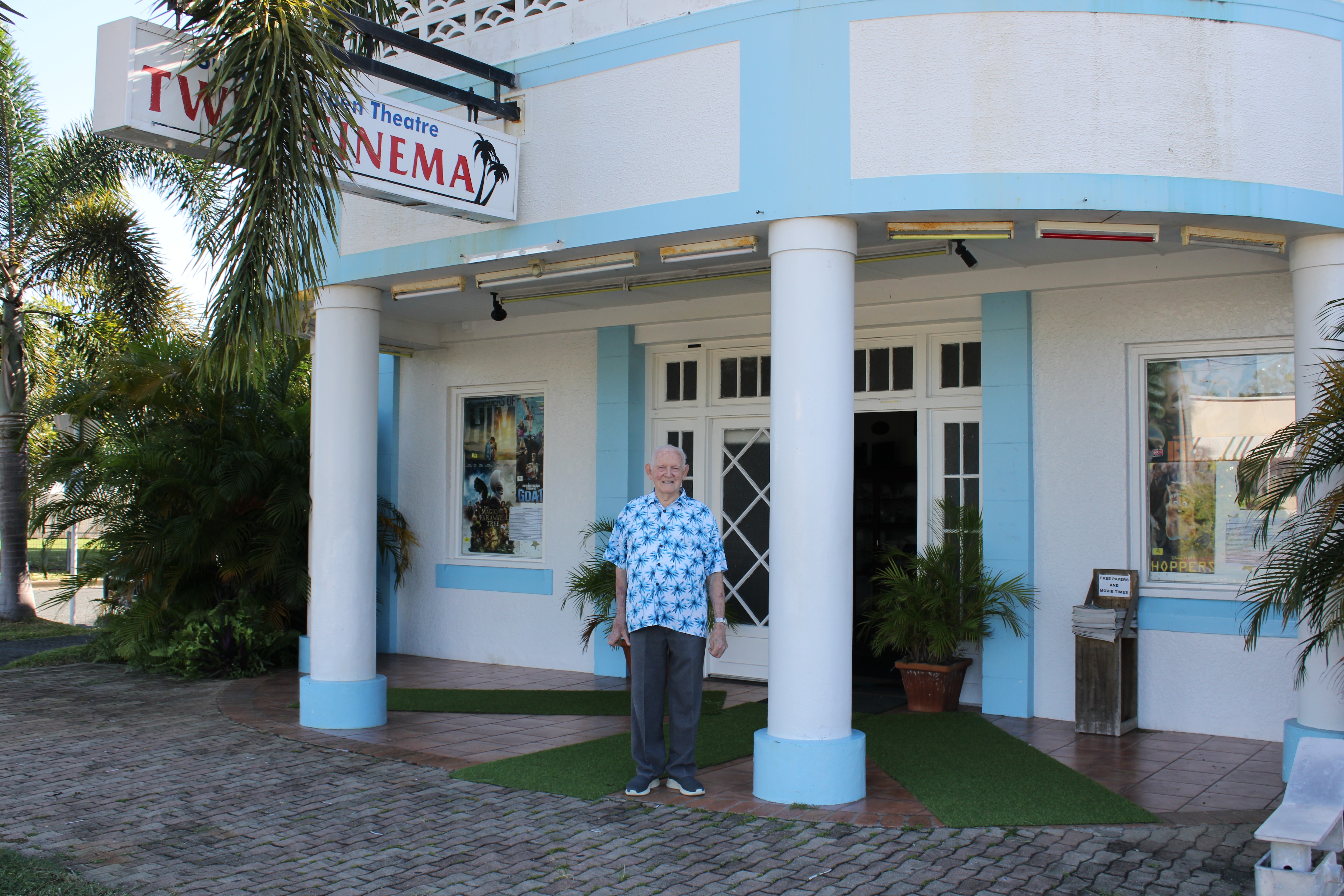 An elderly man in a blue tropical printed shirt standing in front of an old cinema.