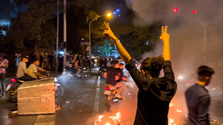 a person in black is seen raising their arms, giving two piece signals, as a street fire burns in front of them