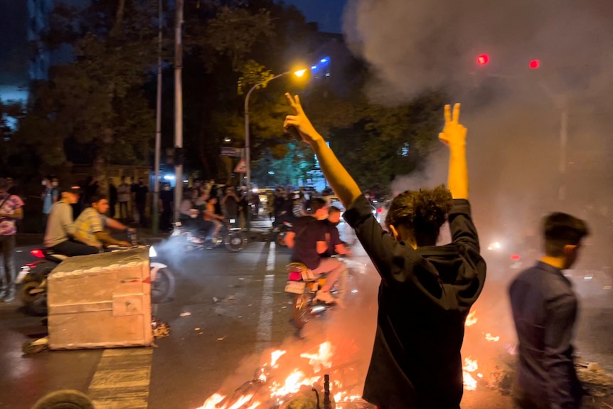 a person in black is seen raising their arms, giving two piece signals, as a street fire burns in front of them