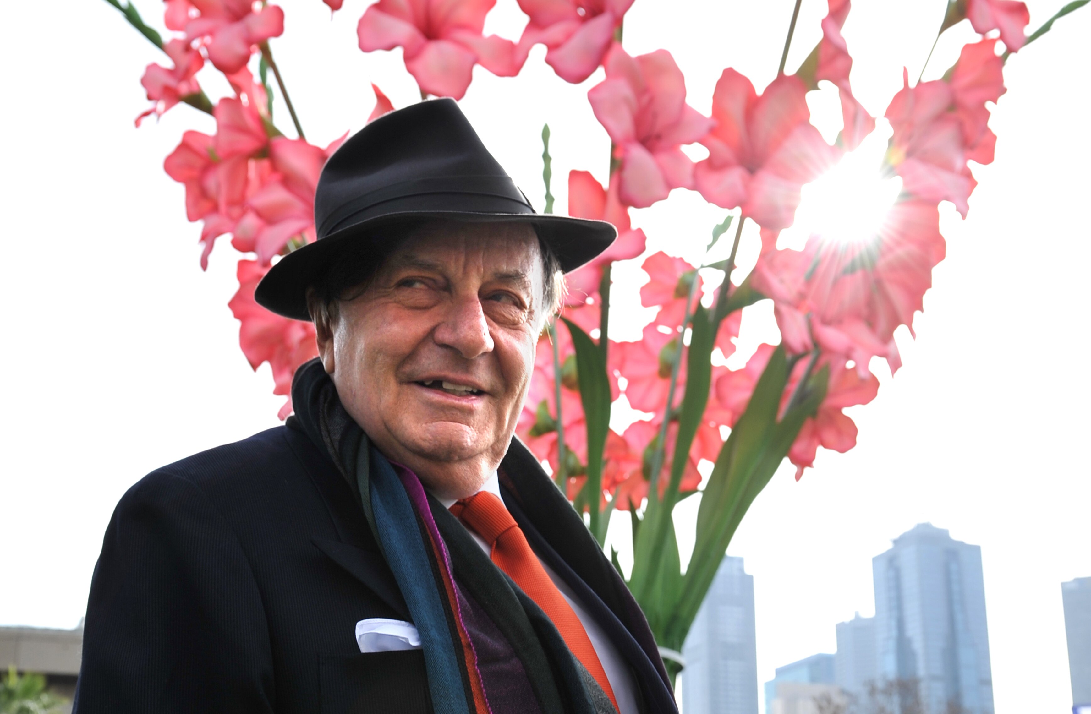 Barry Humphries wears a suit and a hat as he stands in front of a large sculpture of gladioli, which has light softly on it.