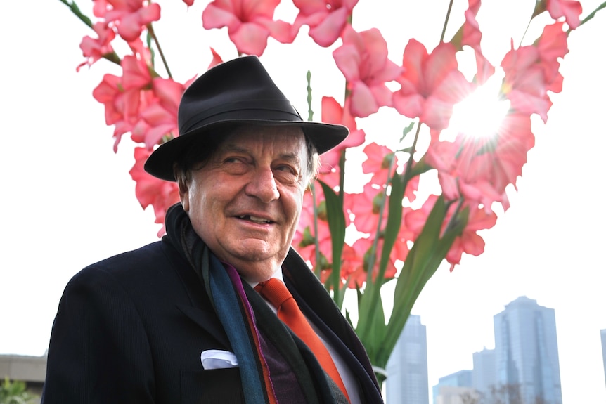 Barry Humphries wears a suit and a hat as he stands in front of a large sculpture of gladioli, which has light softly on it.