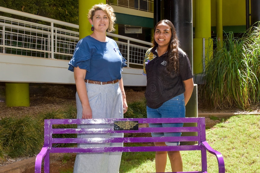 Two women stand behind a bright purple park bench in a university courtyard.