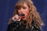 Taylor Swift stands on stage holding microphone wearing black sparkly leotard with long sleeves.