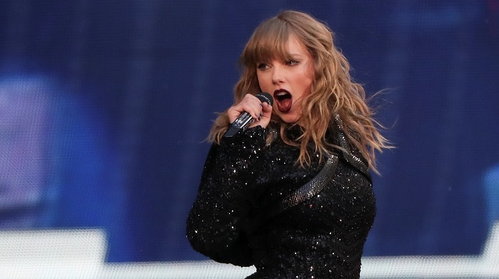 Taylor Swift stands on stage holding microphone wearing black sparkly leotard with long sleeves.