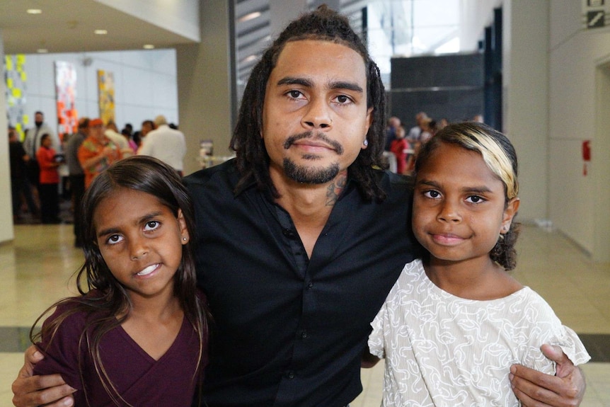 A photo of an Indigenous man holding his two young daughters at an awards ceremony.