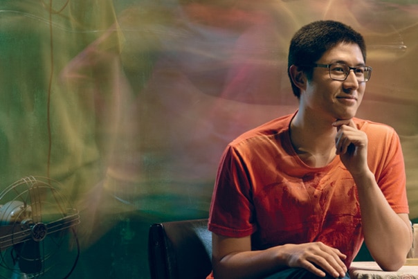 A young man wearing an orange t-shirt and glasses and seated at a table smiles.