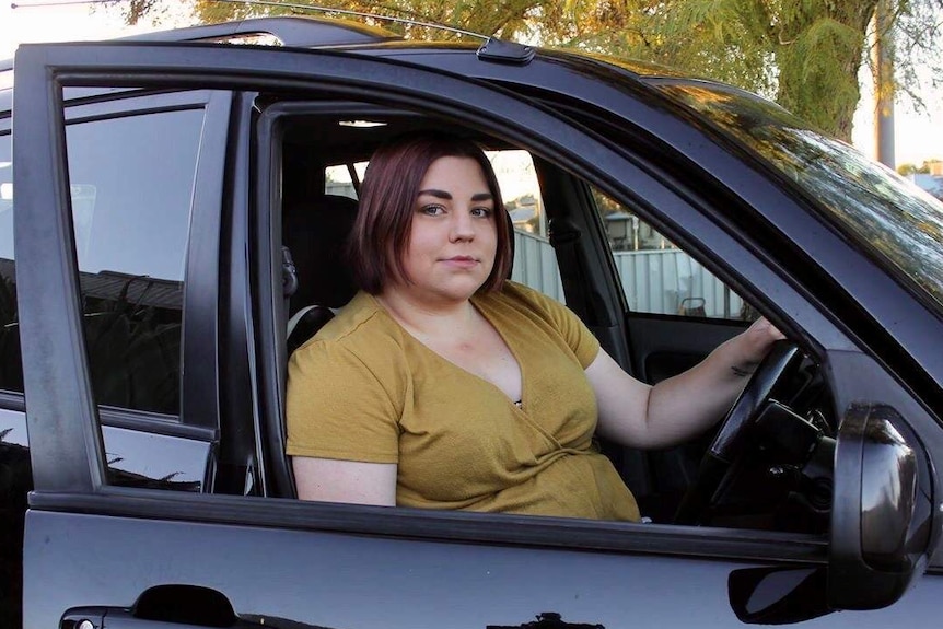 A young woman wearing a mustard yellow top sits in the drivers seat of her shiny black car looking directly at the camera.
