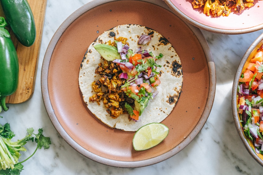 A cauliflower taco on a plate beside a wedge of lime, salsa and jalapeno peppers.