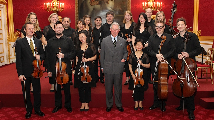 The ACO post in a photo with the Prince of Wales holding their instruments in front of an ornate red wall.