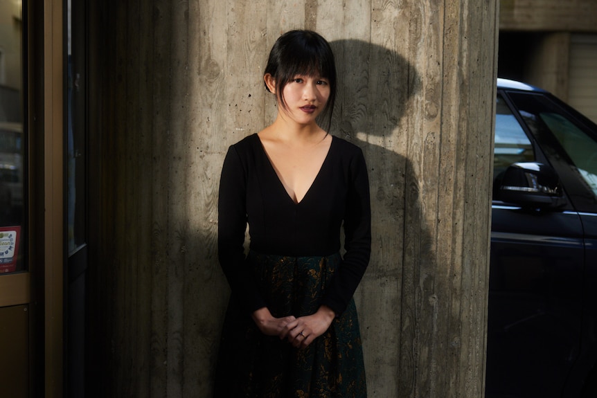 Rebecca F. Kuang wearing all black with hair tied back and hands clasped together in front of her, standing