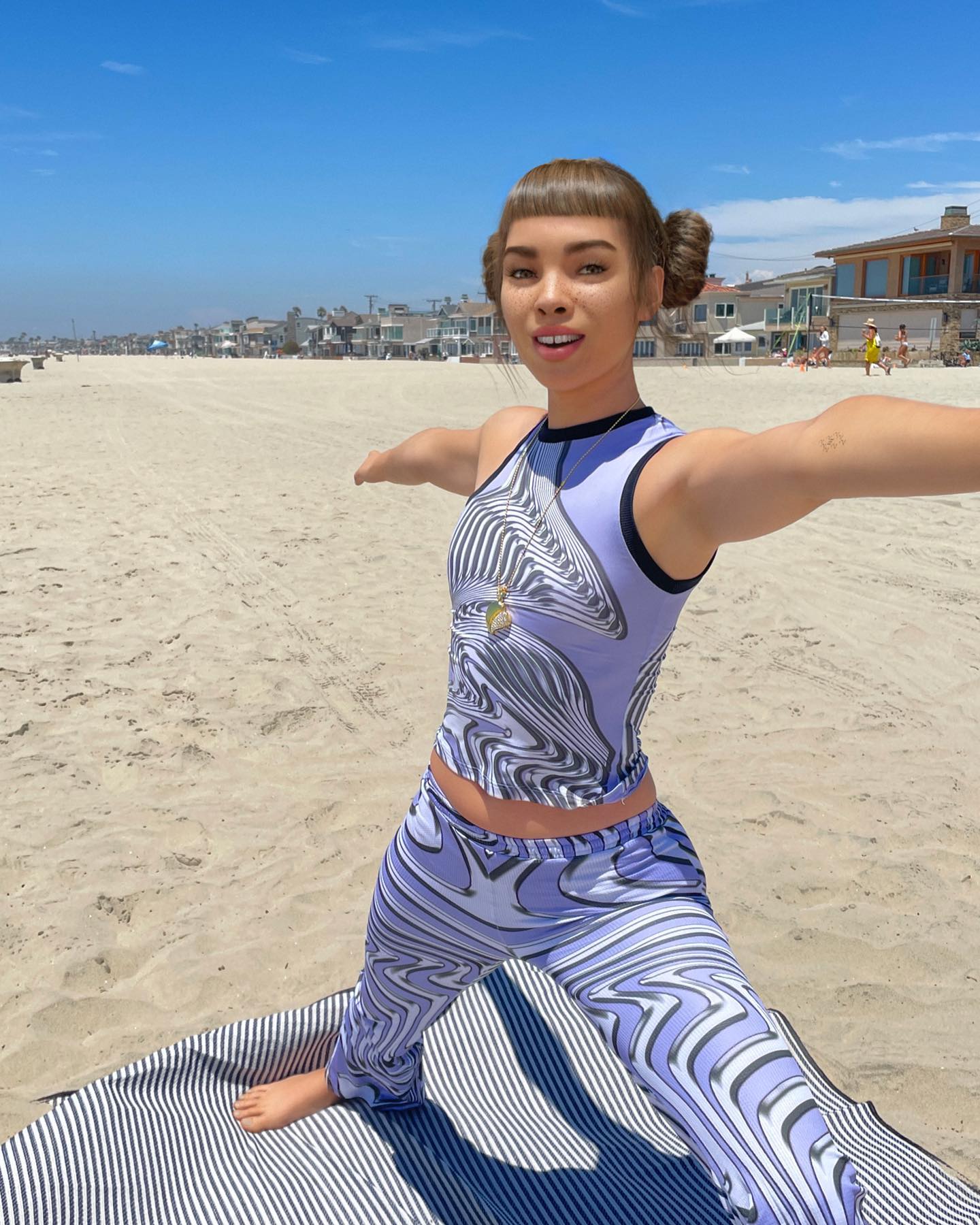 Lil Miquela, dressed in purple, stands in a wide yoga pose on a beach.