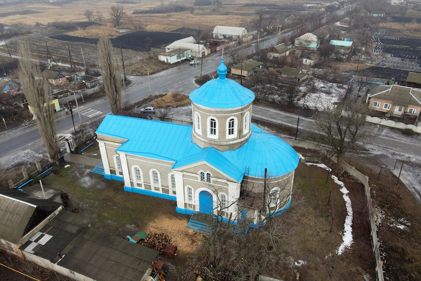 A birds-eye exterior view of a small church with a blue roof, amid a small village.