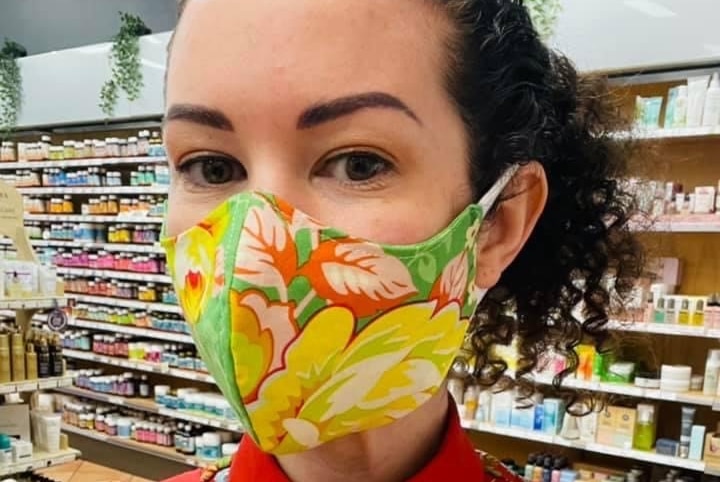 Gen wears a floral mask and stands in her store, wearing an apron.
