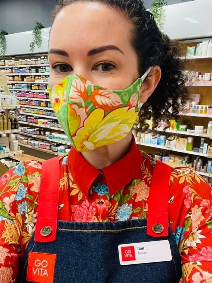 Gen wears a floral mask and stands with an apron in her shop.