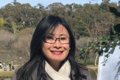 A woman standing near a lake smiling, wearing a jacket and a scarf.