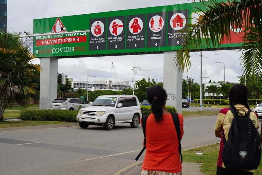Two women prepare to cross a city road in a tropical setting with big green sign straddling road.
