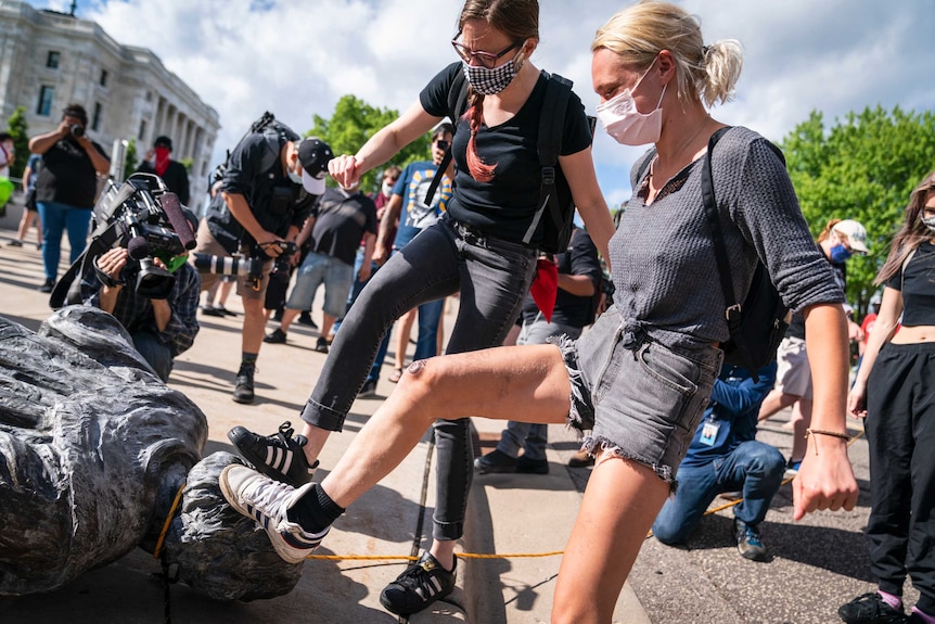 Two women in face masks kick a toppled statue of Christopher Columbus