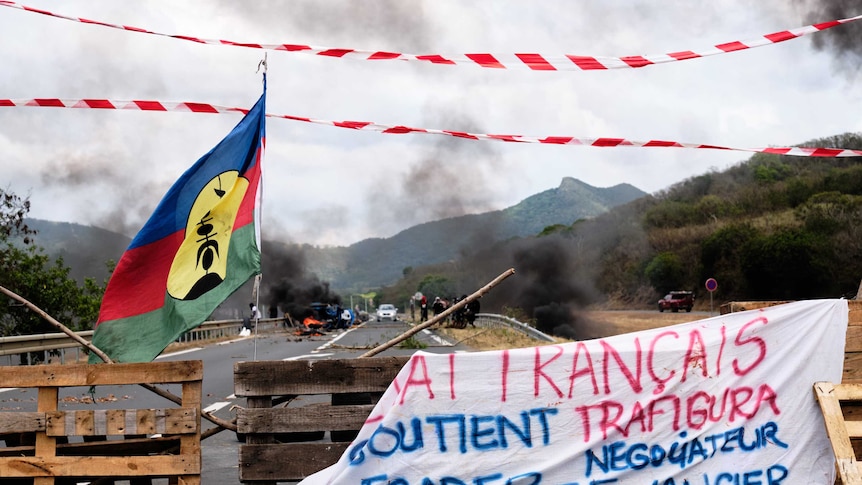 Kanaky flags wave in front of a roadblock, smoke in the distance.