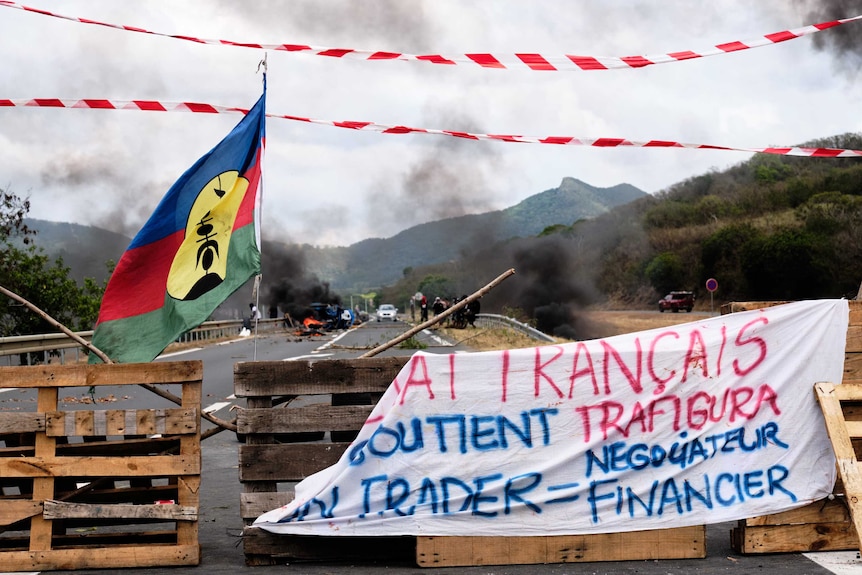 Kanaky flags wave in front of a roadblock, smoke in the distance.