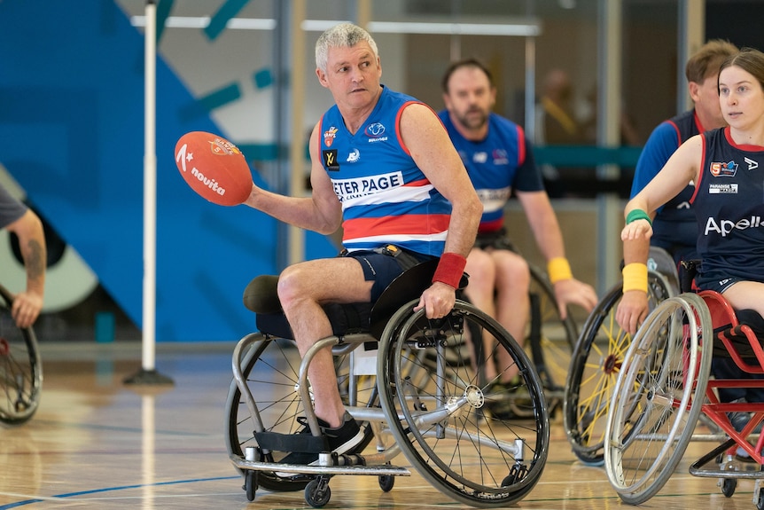 A man in a wheelchair with one leg carrier a red football in one hand while pushing his chair with the other