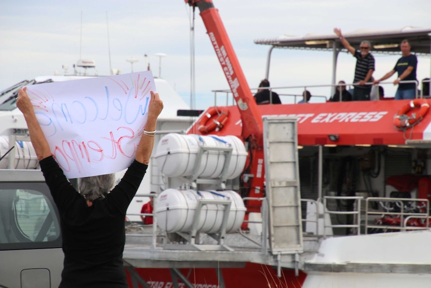 A person holds up a welcome sign as a red and white ferry docks at a wharf.