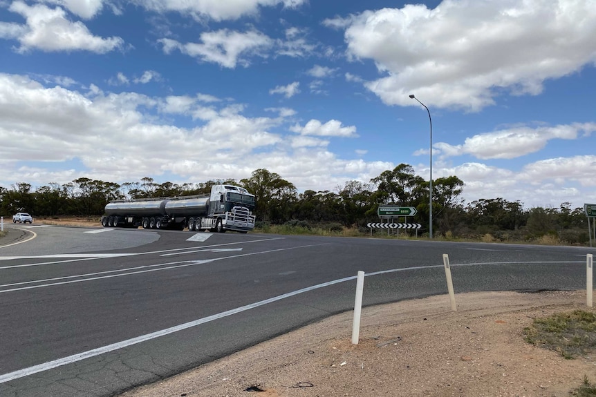 A bend in a country highway road as a large truck waits to turn onto it from a side road.